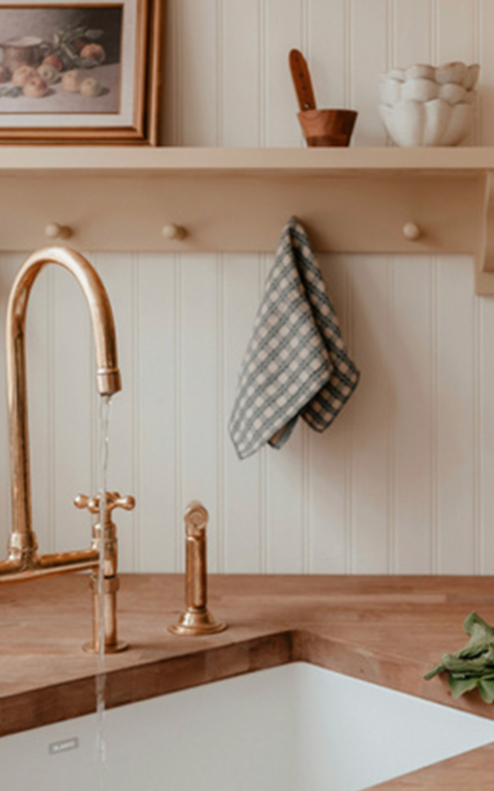 A brass kitchen faucet runs water into a white sink set in a wooden countertop, showcasing thoughtful design. Above, a checkered cloth hangs on a peg rail under a shelf holding a framed painting, a wooden utensil, and a white bowl.
