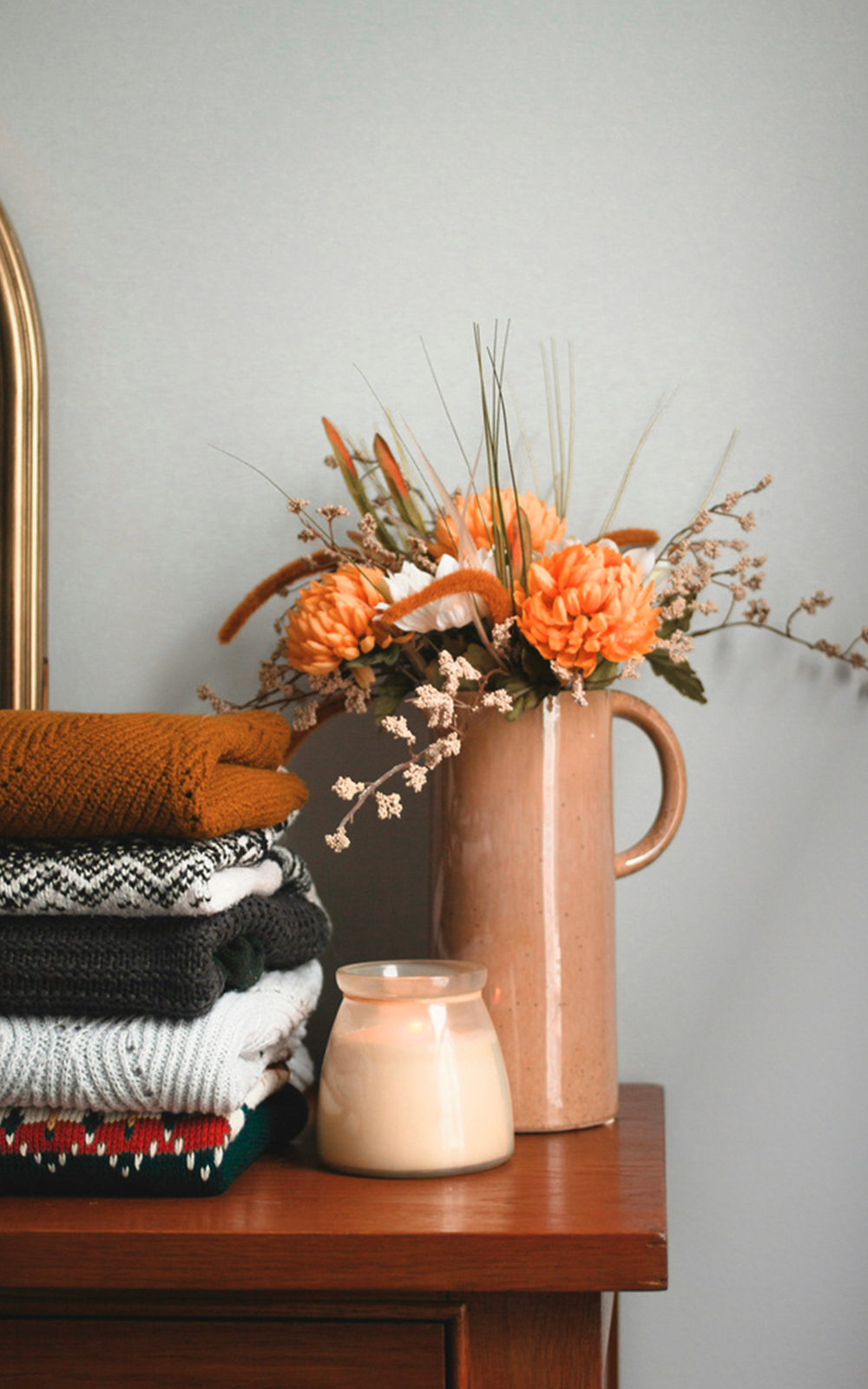 A stack of folded sweaters, a lit cream-colored candle, and a beige vase with orange and white flowers create a cozy design on a wooden surface against a light gray wall.