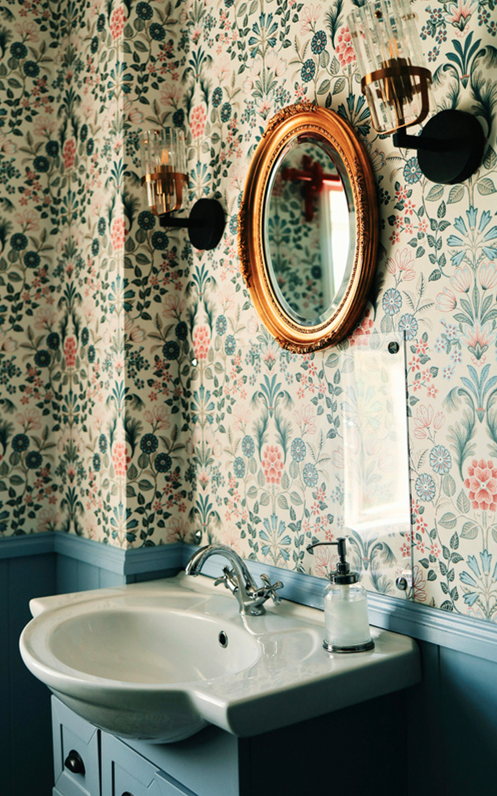A bathroom with floral wallpaper and blue wainscoting showcases thoughtful design, featuring a round gold-framed mirror, two wall sconces, a white sink with a silver faucet, and a soap dispenser on a blue vanity.