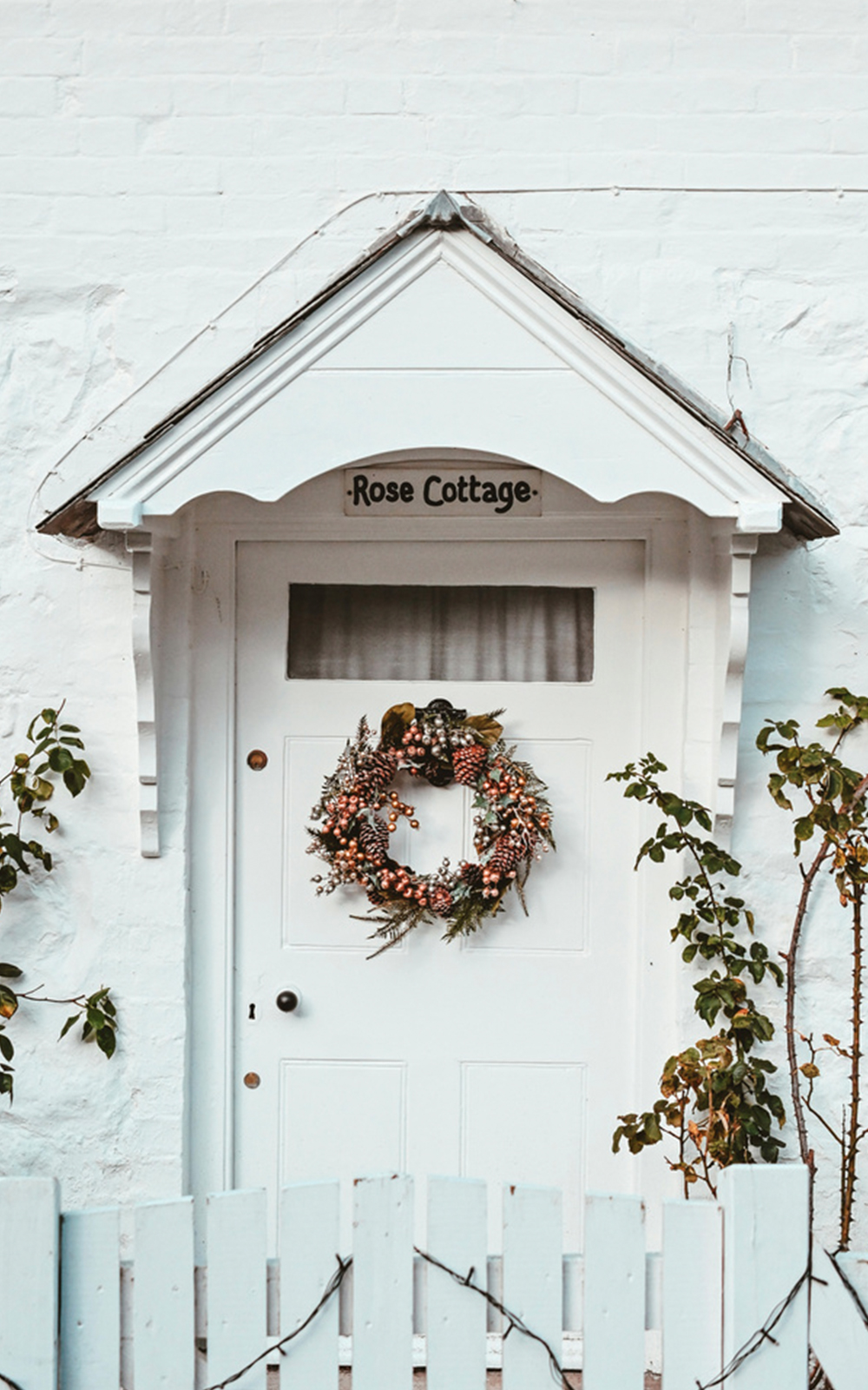 A white front door with a floral wreath and small window, sheltered by a white awning, showcases charming design. A sign above reads "Rose Cottage." Leafy plants flank the door, and a white picket fence frames the entrance.