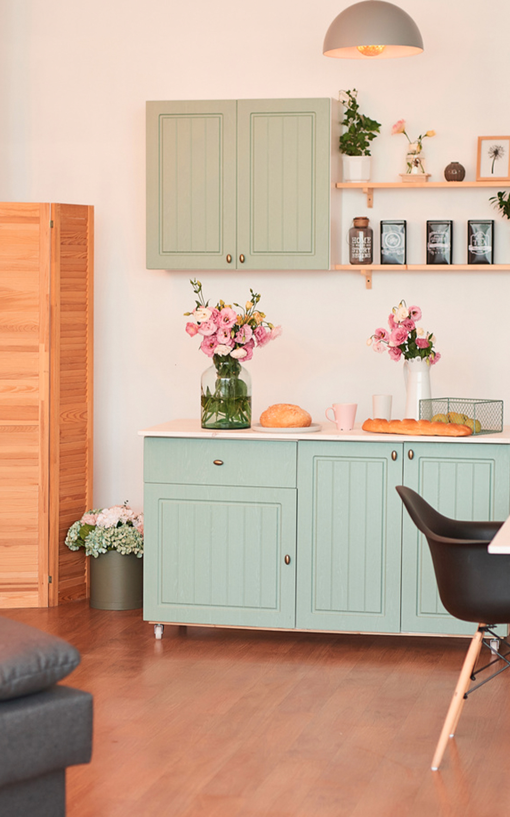 A cozy kitchen with light green cabinets and wooden floor showcases thoughtful design, featuring vases of pink flowers, a loaf of bread, mugs, and decorative items on open shelves. A black chair and a wooden divider complete the inviting space.