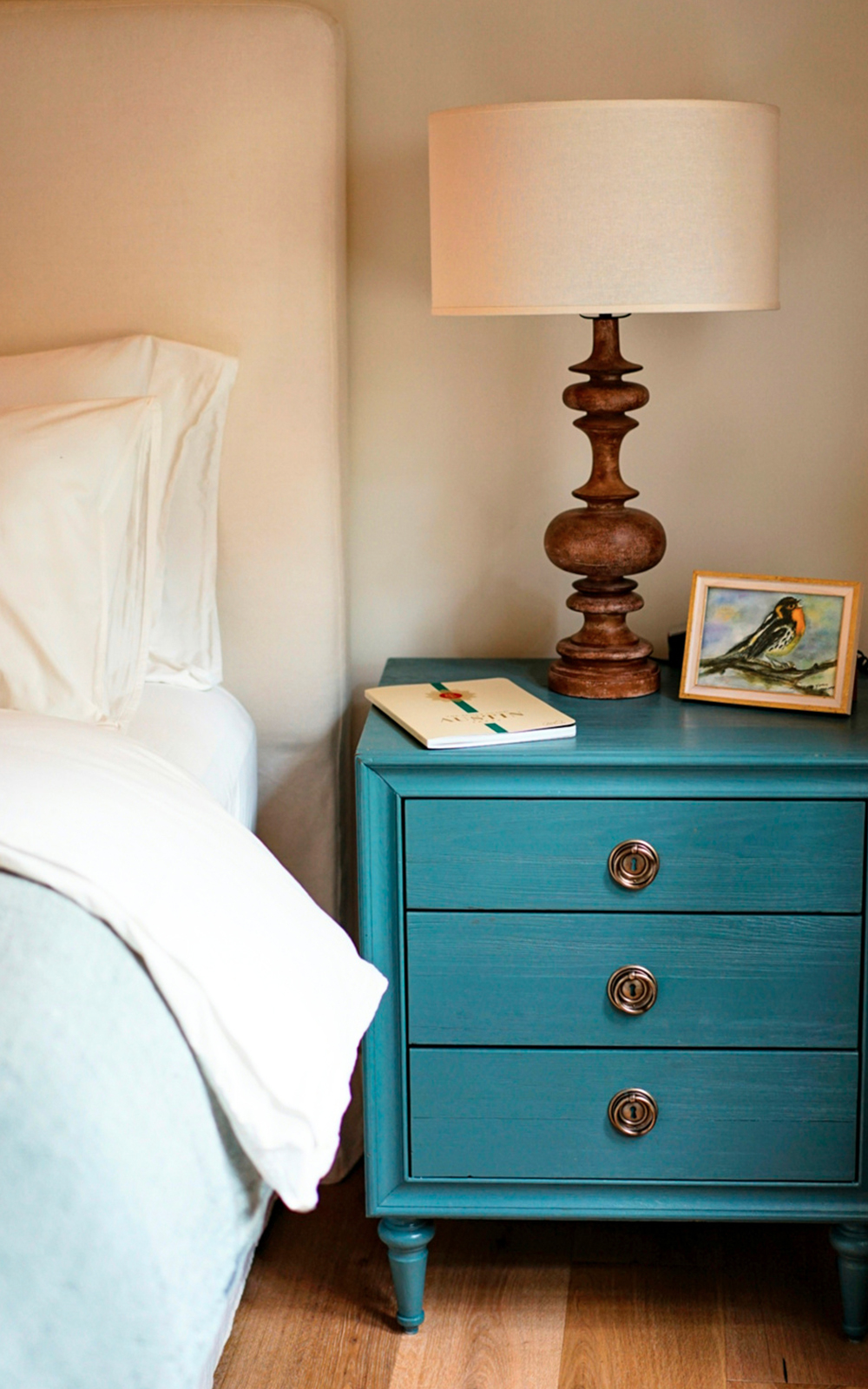 A blue nightstand with three drawers adds a touch of design beside the bed. On top are a wooden lamp with a cream shade, a small painting of a bird in a yellow frame, and an open book or notebook. White bedding is partially visible.