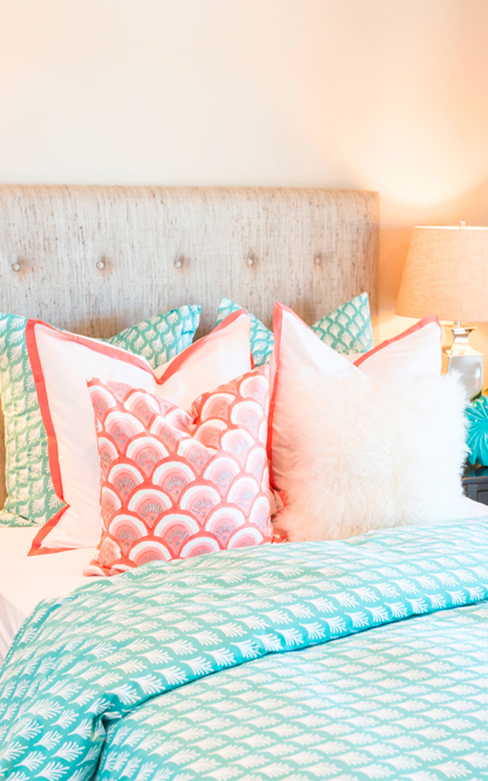 A neatly made bed with a tufted beige headboard showcases thoughtful design, decorated with teal and white pillows, a fluffy white pillow, and a pink patterned pillow. The bed features a teal and white patterned comforter. A lamp sits on the nightstand.