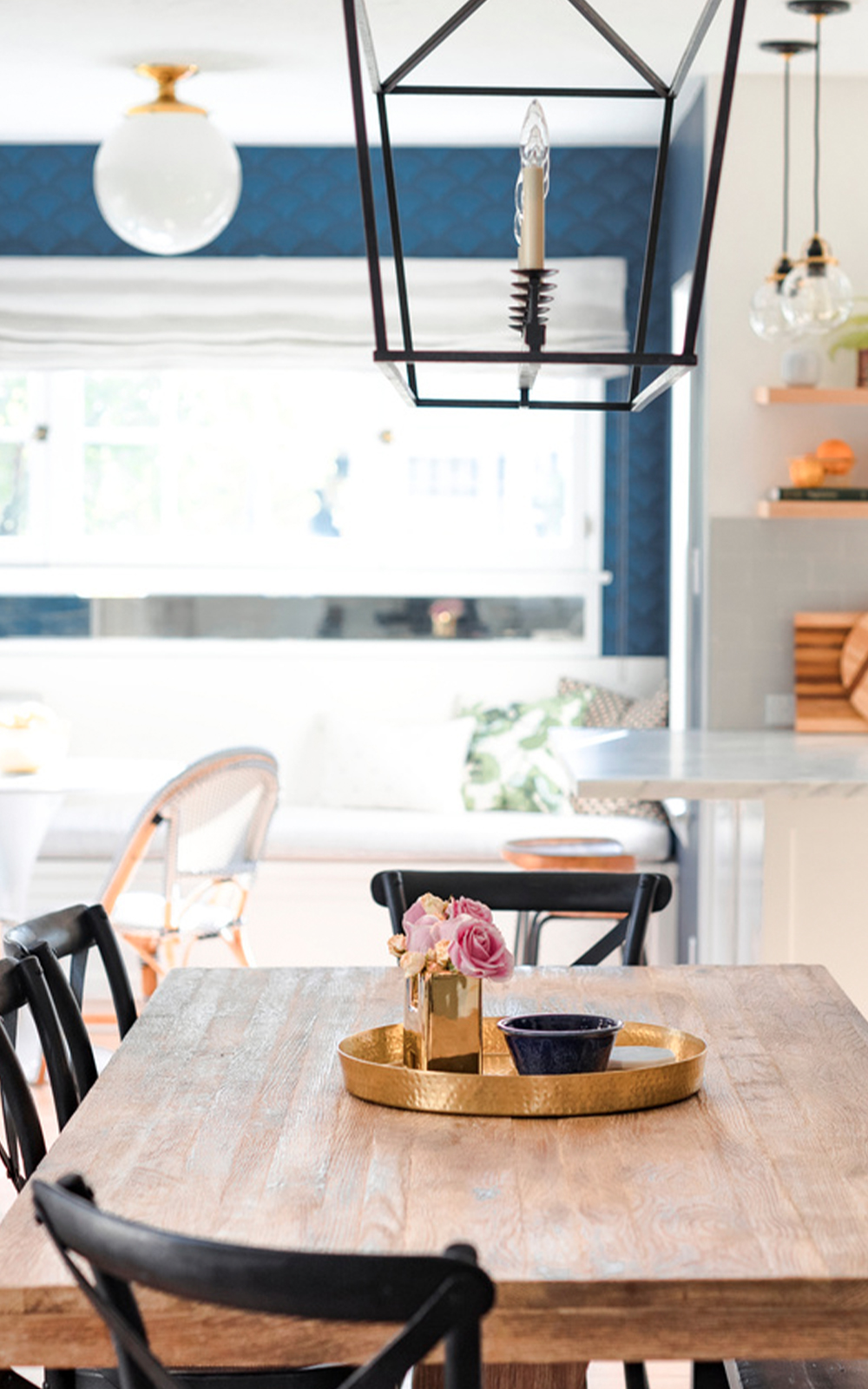 A bright dining area with a wooden table, black chairs, and a gold tray holding flowers and a bowl. A modern lantern-style light adds to the design, while a window seat and blue accent wall are visible in the background.