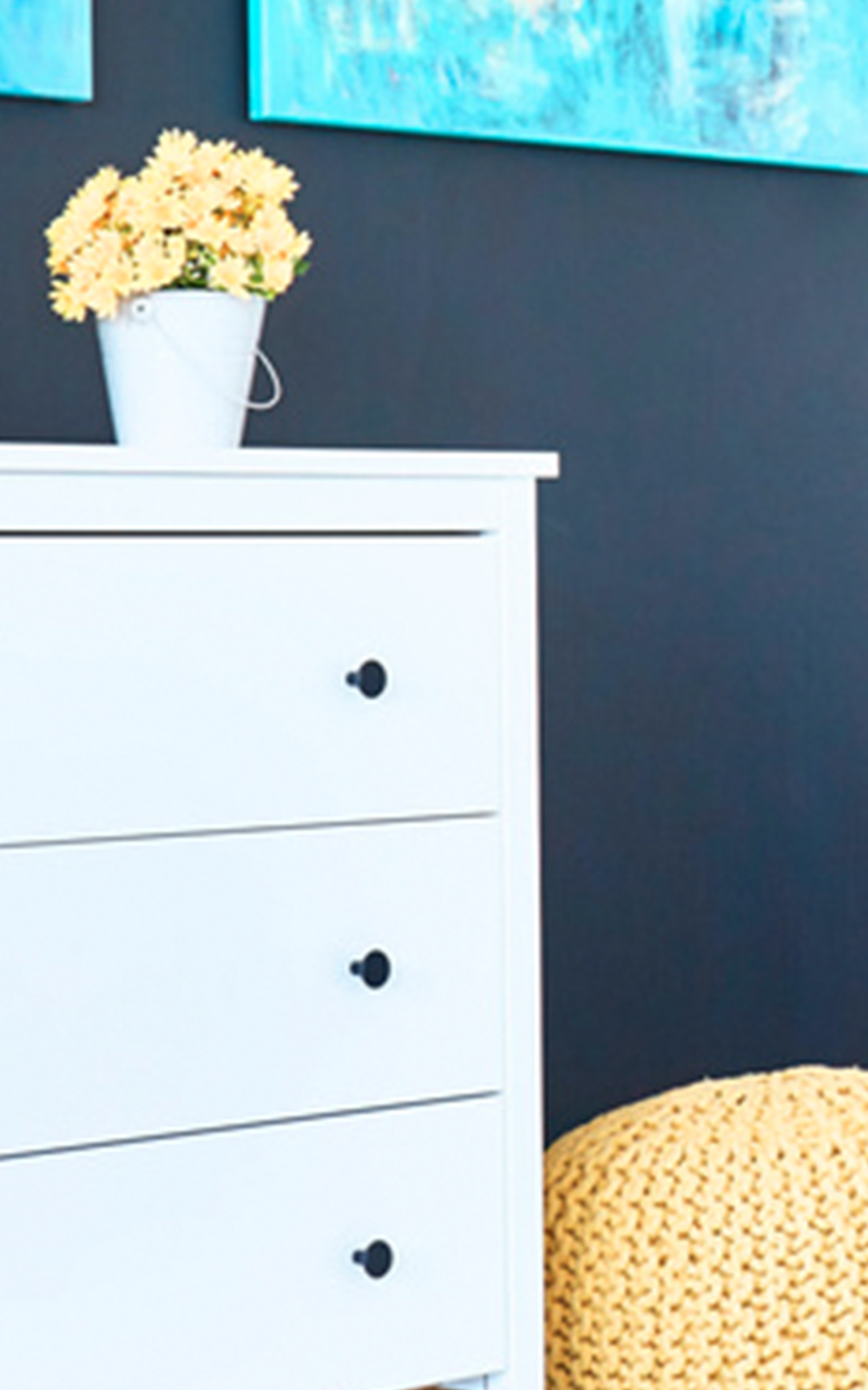 A white dresser with black knobs stands beside a yellow knitted pouf, showcasing thoughtful design. A white pot of yellow flowers sits on top, while a blue abstract painting hangs on a dark wall in the background.