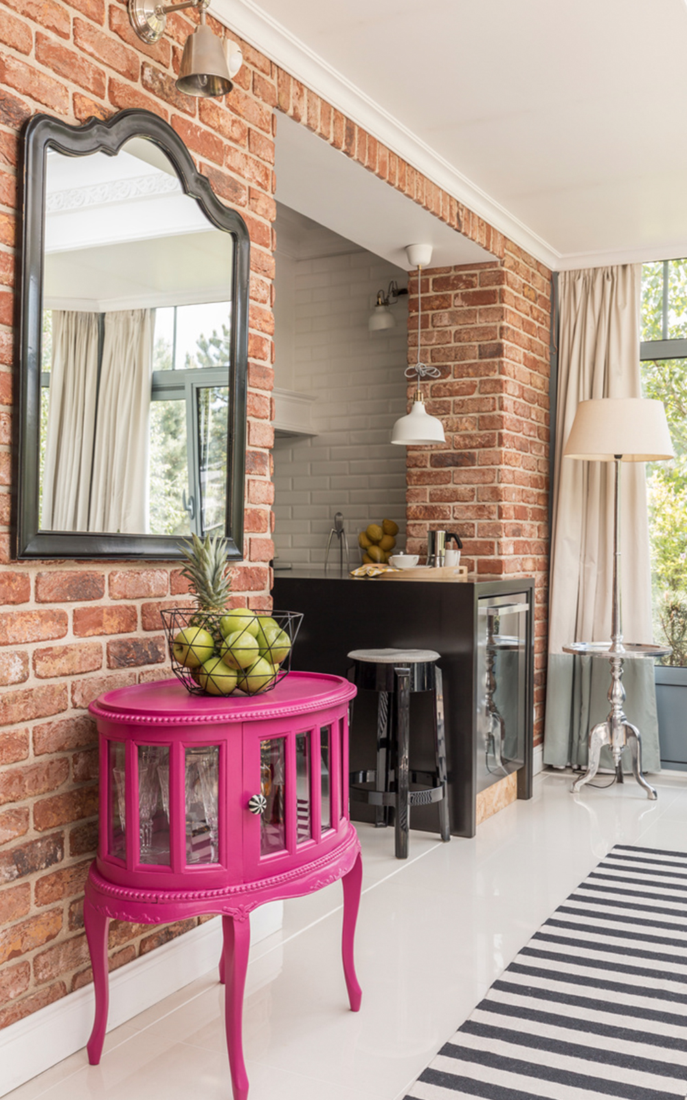 A modern kitchen with exposed brick walls features thoughtful design elements like a black-framed mirror, a bright pink side table with green apples, a black and white striped rug, and large windows with cream curtains.