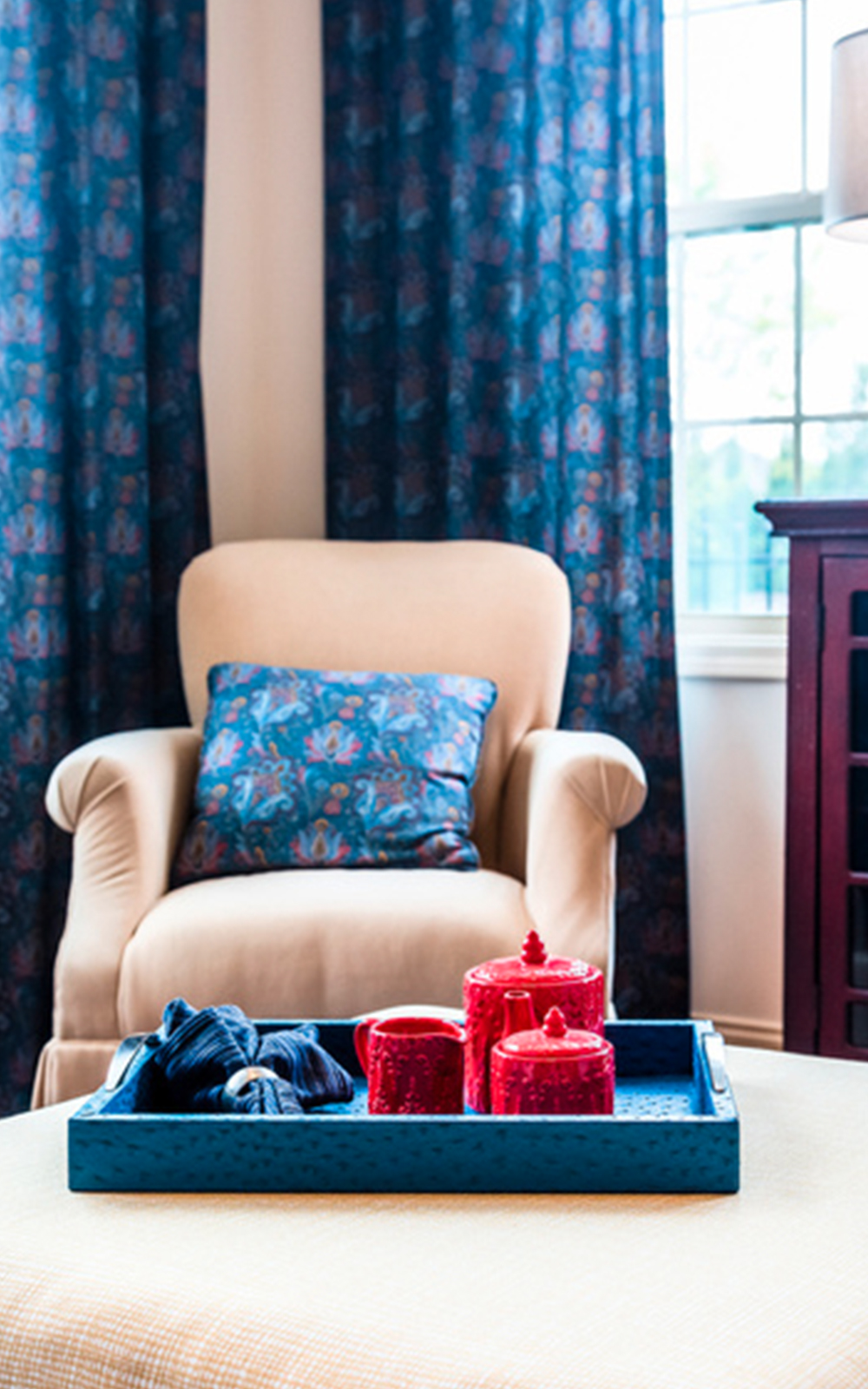 A cozy beige armchair with a blue cushion sits by a window adorned with blue patterned curtains, showcasing thoughtful design. In the foreground, a blue tray with red cups and a cloth rests on a light yellow ottoman.