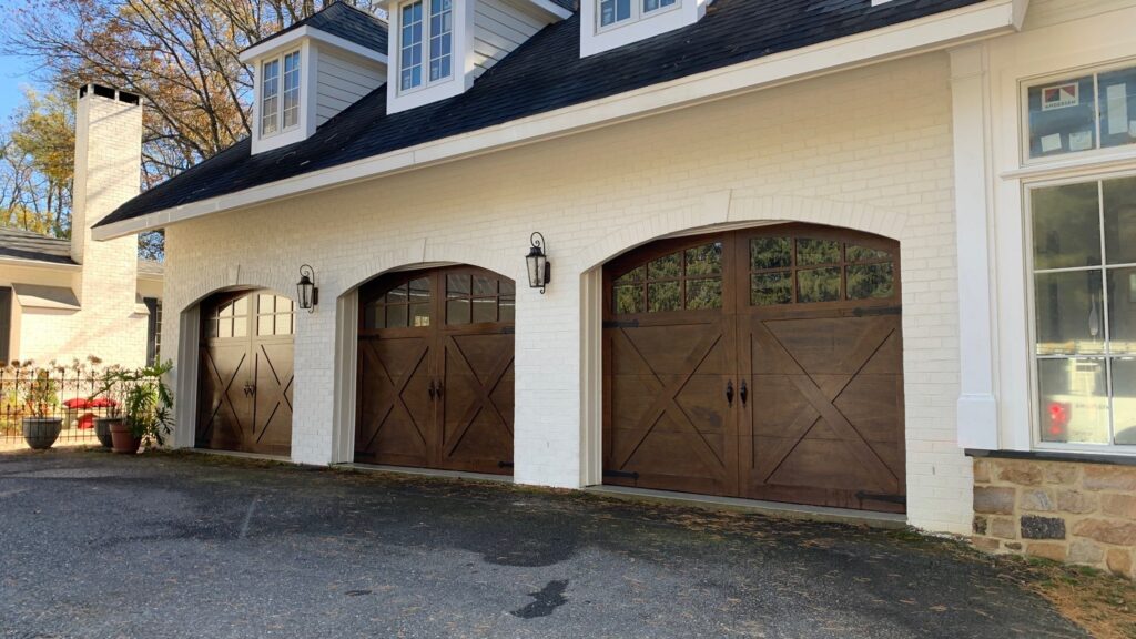 Three wooden garage doors with decorative black hardware are set in a white brick exterior of a house. Two lantern lights are mounted between the doors, and a paved driveway is in front.