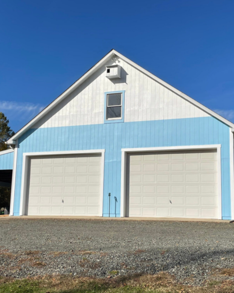 A light blue and white garage with two closed white doors, a small window above, and a clear blue sky in the background. The ground in front is gravel.