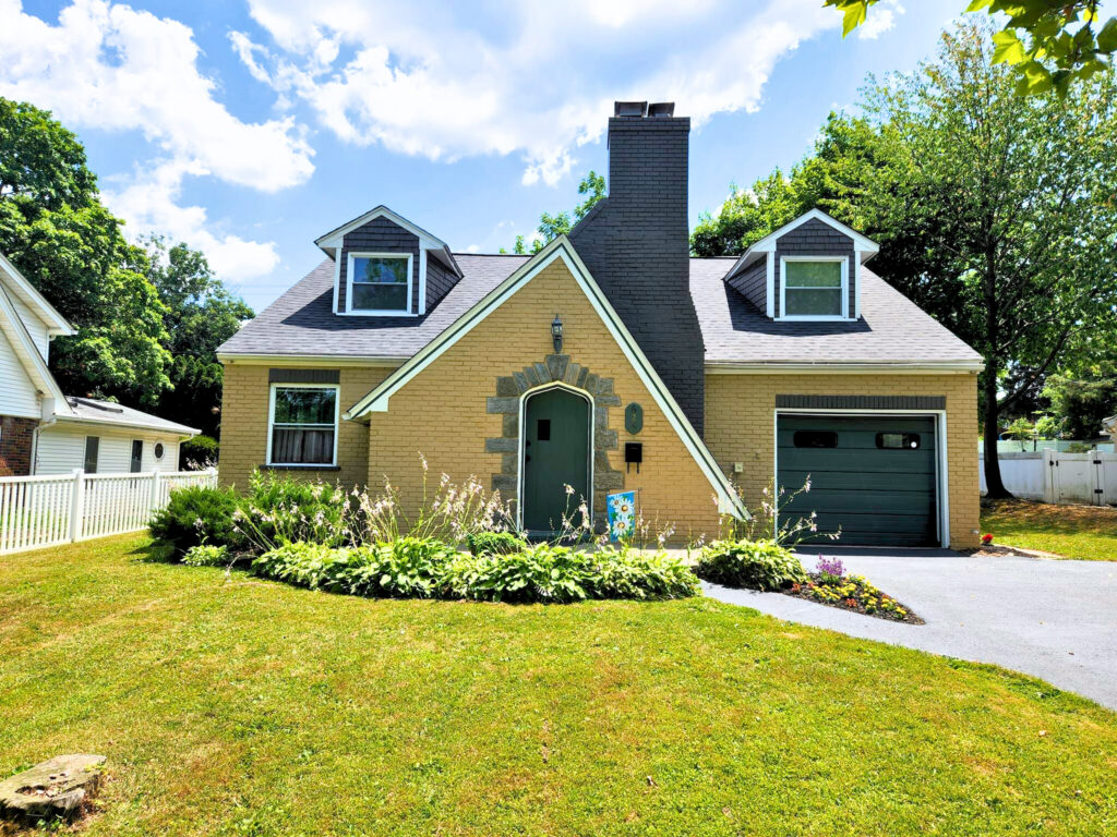 A tan brick house with a steep roof, two dormer windows, a central front door, and a single-car garage, surrounded by green grass, plants, and trees under a partly cloudy sky.