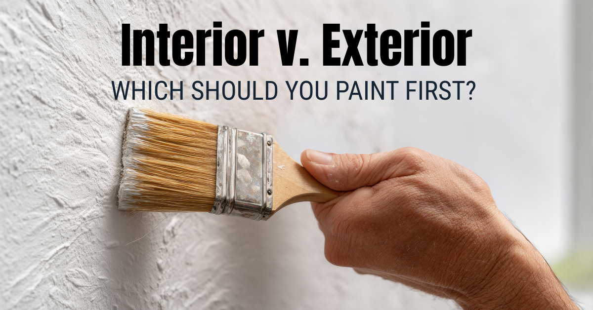 A hand holds a paintbrush, applying white paint to a textured wall. Text above reads: "Interior v. Exterior: Which Should You Paint First?.