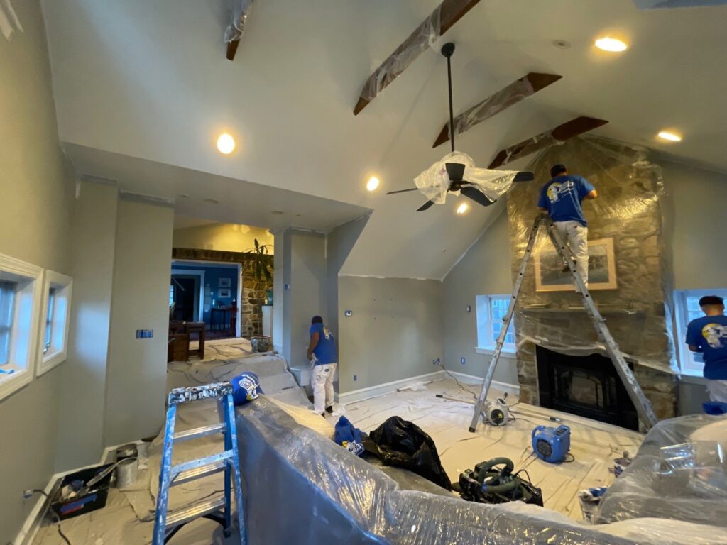 Three workers in blue shirts and white pants are painting and preparing a living room. Furniture and floors are covered with plastic, and ladders and tools are scattered around. One worker is on a ladder near a stone fireplace.