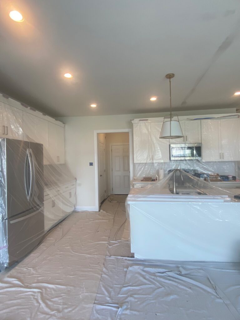 A kitchen with appliances, cabinets, and countertops covered in plastic sheeting for protection. The floor is also covered, and recessed ceiling lights and a pendant light are visible. The space appears to be prepared for painting or renovation.