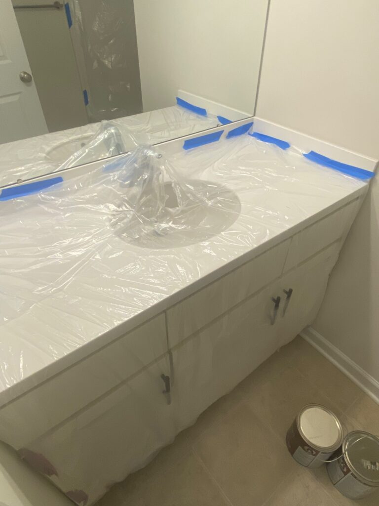 A bathroom vanity and sink covered in plastic and blue painter’s tape, with paint cans on the floor nearby, preparing the area for painting or renovation.