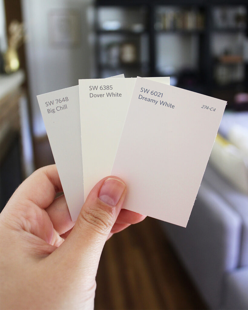 A hand holds three paint swatches labeled SW 7648 Big Chill, SW 6385 Dover White, and SW 6021 Dreamy White, with a blurred living room in the background.