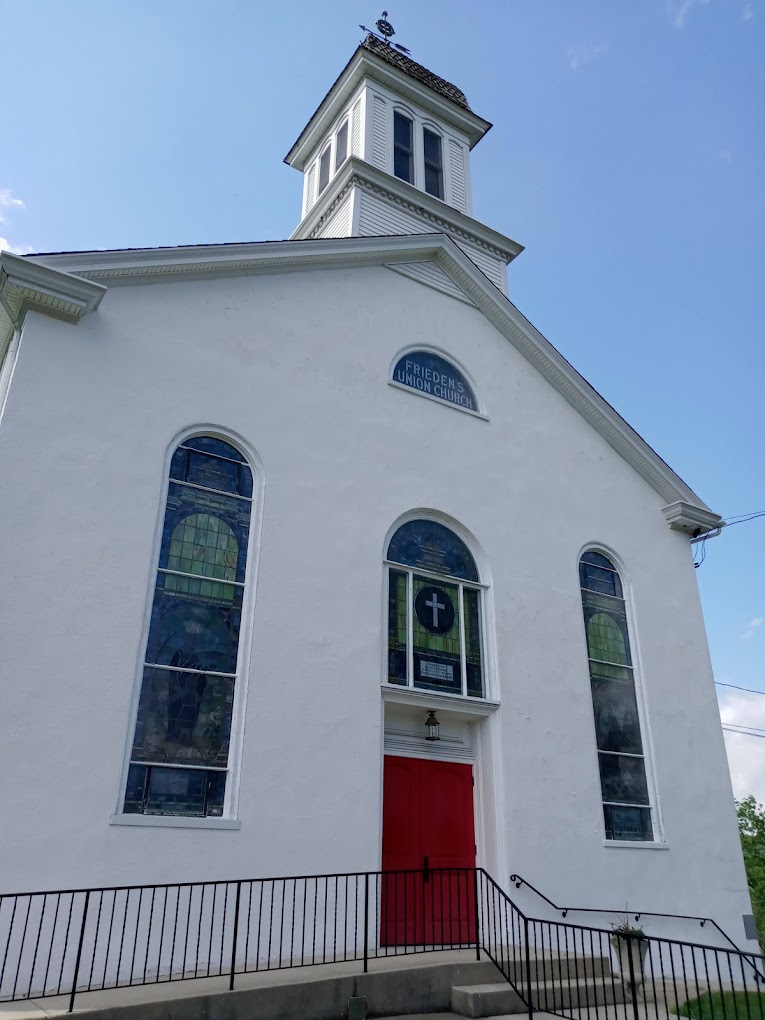 A white church with tall arched stained glass windows, a red double door, and a bell tower against a partly cloudy blue sky. The sign above the door reads "BRETHREN UNION CHURCH." A railing lines the entrance steps.
