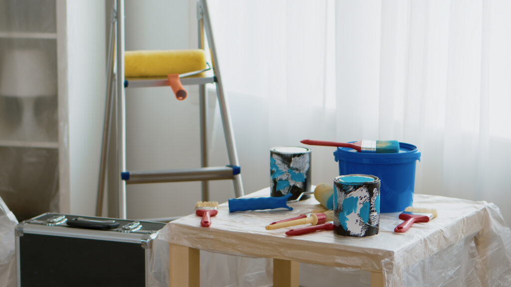 A small table covered with plastic holds paint cans, brushes, and a blue bucket. A ladder with a paint roller leaning on it stands nearby in a bright room with sunlight coming through sheer curtains.