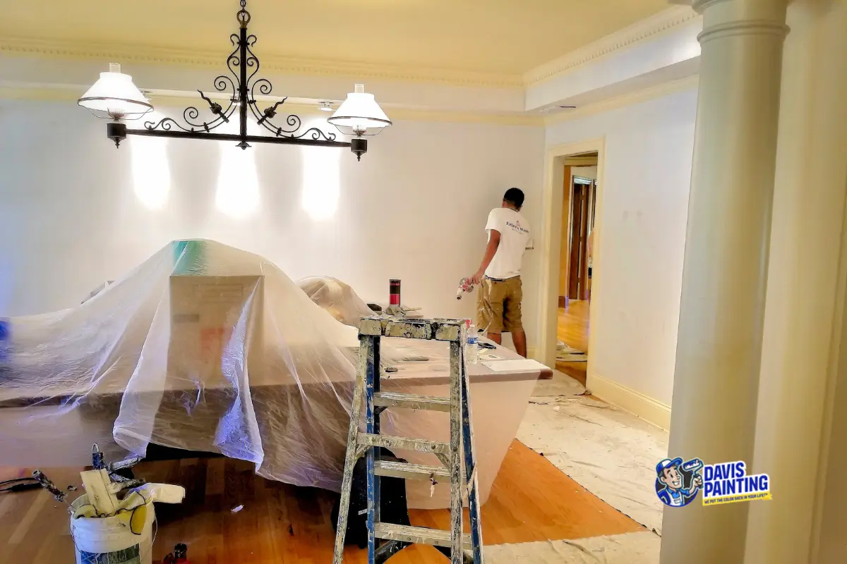 A person performs interior painting on a large white wall in a room. Furniture and a pool table are covered with plastic sheets, while a ladder and painting supplies sit in the foreground. The "Davis Painting" logo appears in the lower right corner.