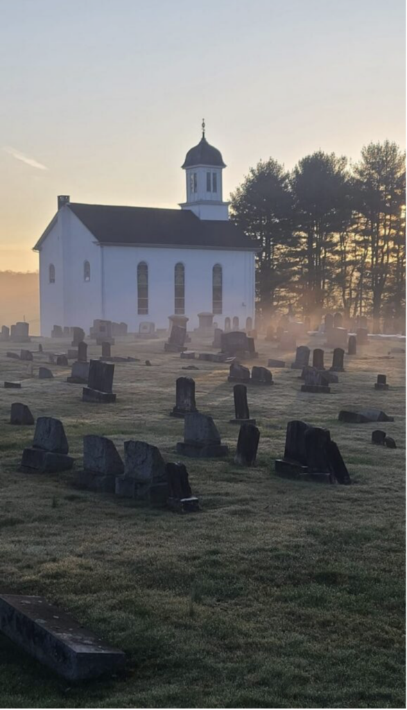 A white church sits behind a foggy graveyard at sunrise, with gravestones scattered across dewy grass and tall trees silhouetted in the background.