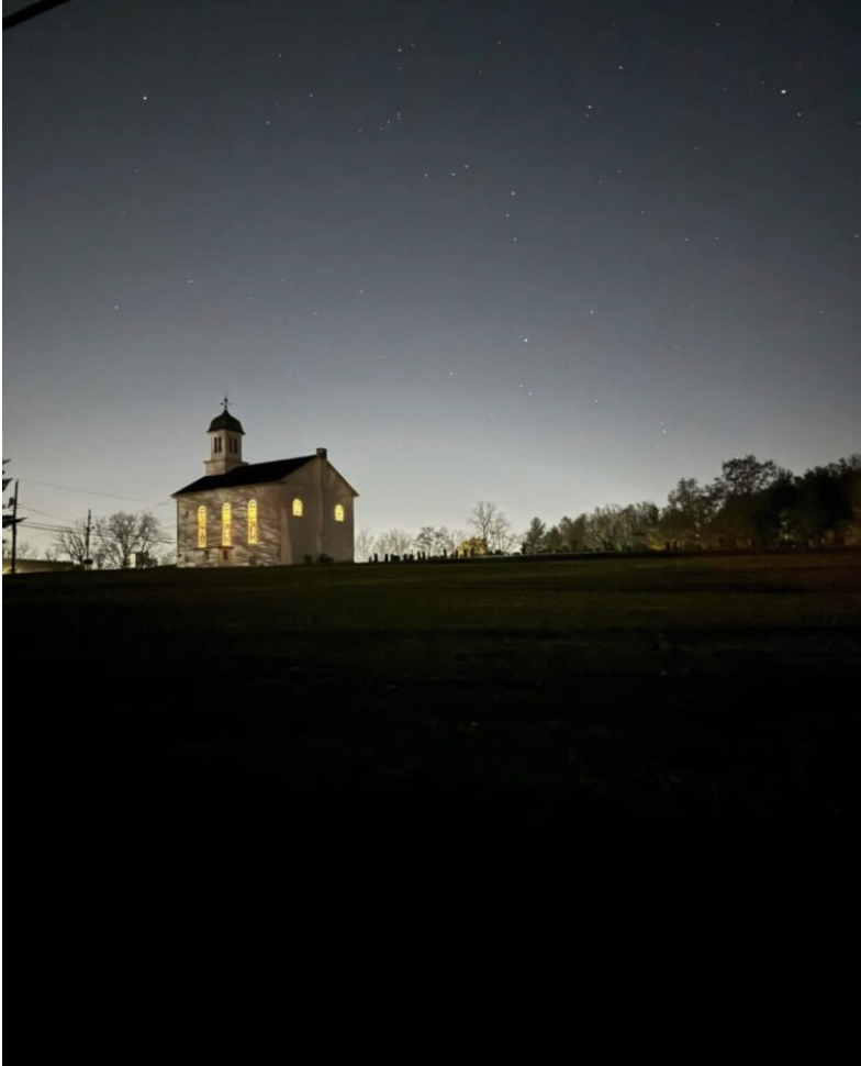 A small church with glowing windows stands on a hill under a starry night sky, surrounded by trees and open land.