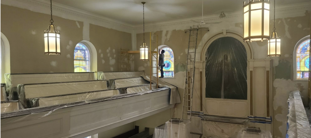 A church interior under renovation with stained glass windows, covered pews, ladders, scaffolding, and plastic sheets protecting surfaces; large hanging lights illuminate the space.