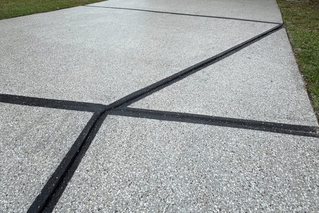 A close-up view of a concrete driveway with black sealant filling the expansion joints, and grass visible on the right edge.
