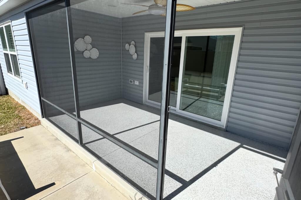 A screened-in porch with light gray flooring, gray siding, sliding glass door, ceiling fan, and white wall decor. Sunlight casts shadows on the enclosed space.