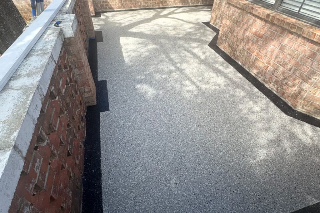 A sunlit patio with speckled light gray flooring and a black border, enclosed by brick walls and a metal railing. Shadows from a nearby tree are visible on the ground.