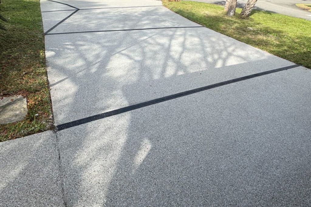 A concrete sidewalk with black expansion joints, casting tree branch shadows on its surface. Grass borders both sides of the walkway.
