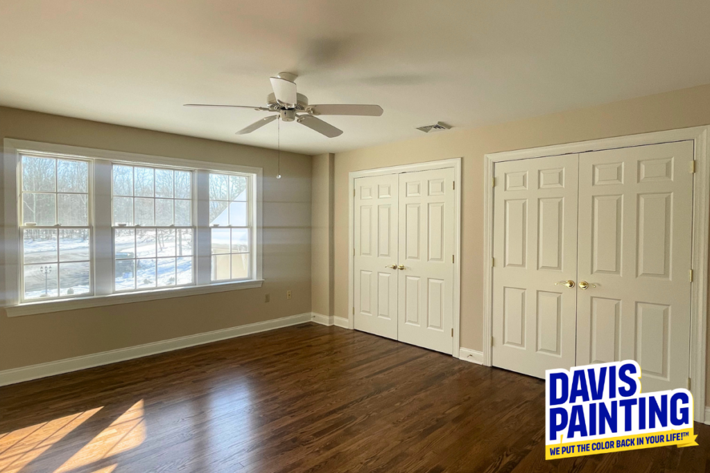 Bright, empty room with tan walls, wood floors, large window, ceiling fan, and white double closet doors. “Davis Painting” logo in bottom right corner.