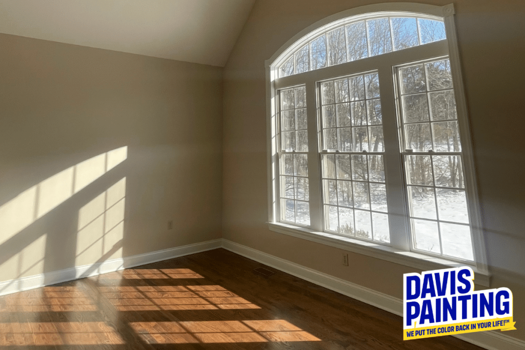 Sunlight streams through a large arched window into an empty, freshly painted room with hardwood floors. “Davis Painting” logo is in the bottom right corner.