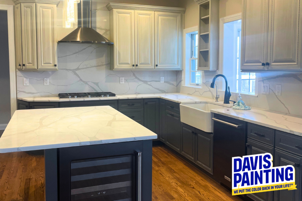 Modern kitchen with light and dark cabinetry, marble countertops, farmhouse sink, stainless steel appliances, and hardwood floor. “Davis Painting” logo in bottom right corner. Sunlight streams in through windows.