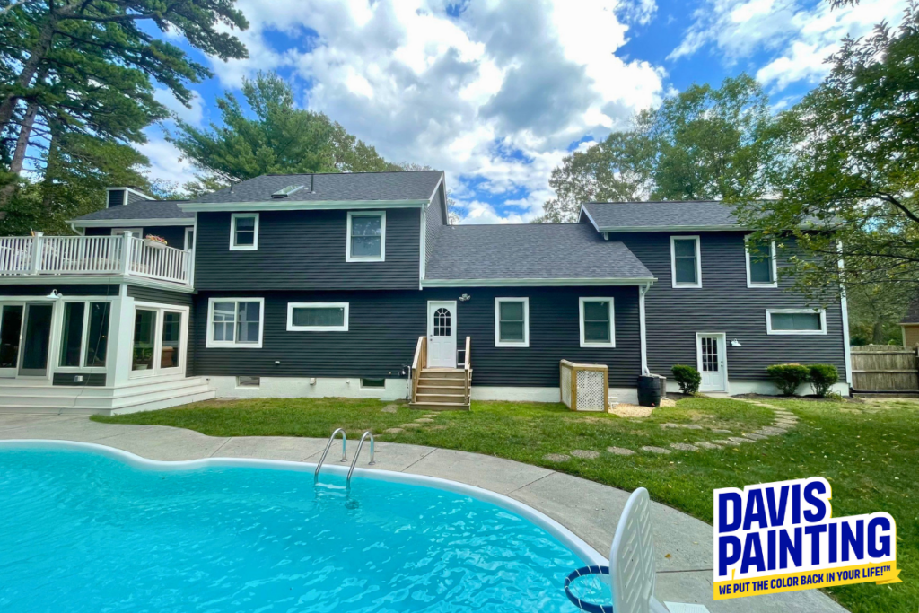 A modern dark-colored house with a pool in the foreground, surrounded by trees. The sky is partly cloudy. A “Davis Painting” logo appears in the bottom right corner, with the slogan, “We put the color back in your life!”.