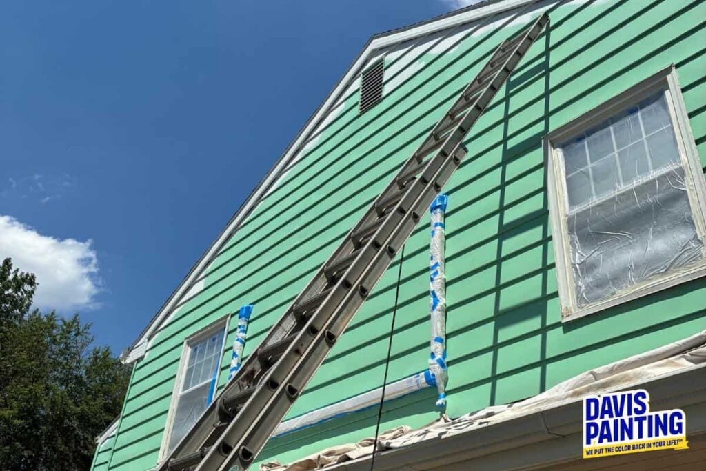 Green house with a tall ladder against its side, windows covered for protection, and the Davis Painting logo in the lower right corner. The sky is clear with some clouds. Exterior Paint