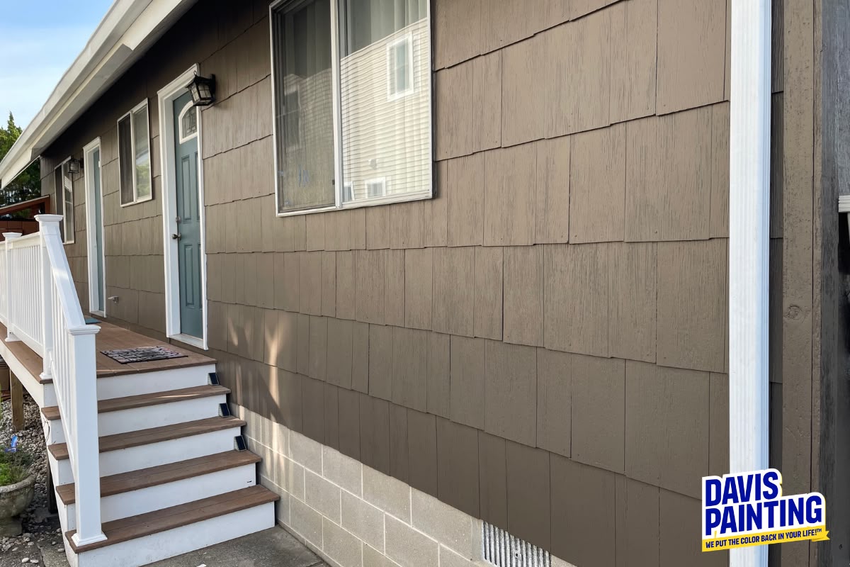A house with brown painted siding, a light blue door, and white trim. Steps with a white railing lead up to the entrance. As a trusted Commercial Painting Contractor, “DAVIS PAINTING” ensures top-quality finishes—logo visible in the bottom right corner.