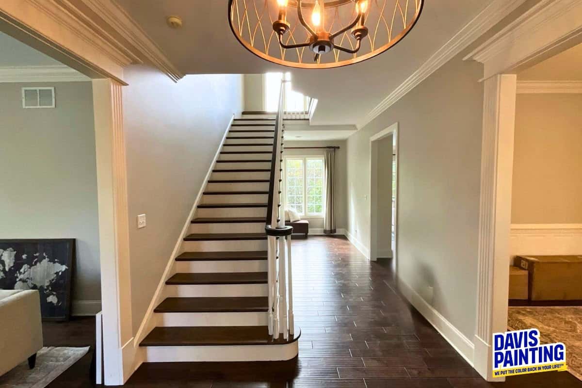 A bright hallway with a wooden staircase, modern chandelier, dark wood floors, beige walls, and a “Davis Painting” logo in the lower right corner highlights the quality of Residential Interior Painting Davis. Light streams in from windows at the end of the hall, showcasing the impeccable craftsmanship you can expect from trusted Interior Painting Services Pennsylvania.