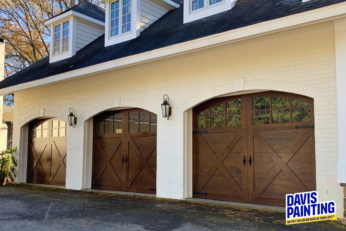 Three wooden garage doors with decorative windows on a white brick house showcase the beauty of fresh exterior paint. Two black lanterns are mounted between the doors, highlighting the home’s inviting façade. A “Davis Painting” logo is in the bottom right corner.